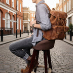 Person sitting on a stool wearing a brown leather backpack against a dark textured wall.