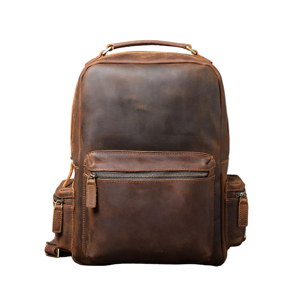 A brown distressed leather backpack with a top handle, front zippered pocket, and side pockets, displayed on a rustic wooden surface against a gray concrete wall.