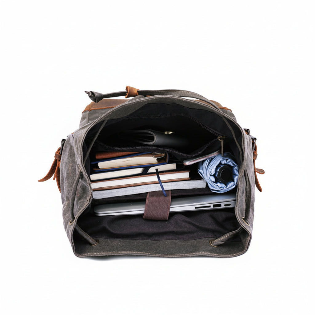 A dark gray canvas crossbody bag shown open from above, filled with notebooks, a laptop, and a rolled blue item. The bag has brown leather trim and a black interior.