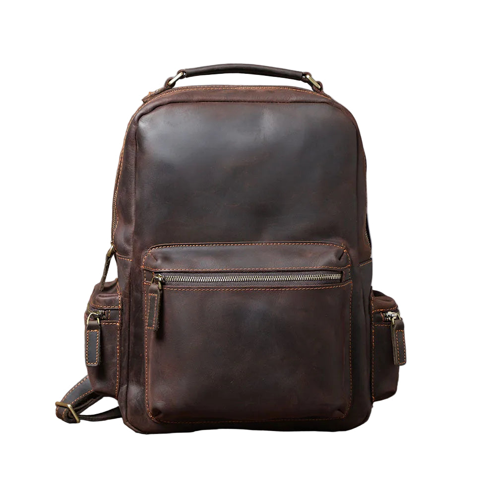 A dark brown distressed leather backpack with multiple zippered compartments and side pockets, displayed on a rustic wooden surface against a gray concrete wall.
