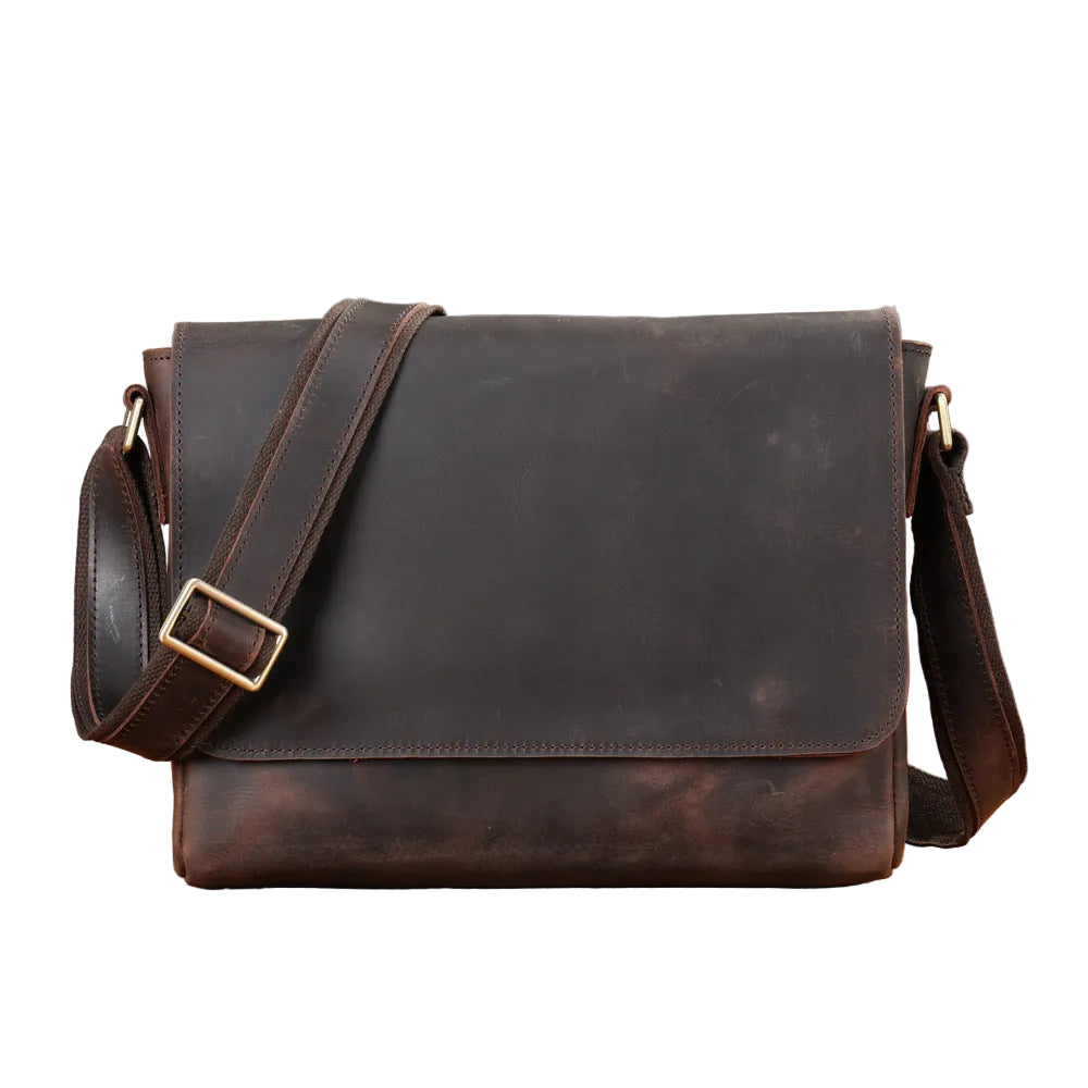 A dark brown distressed leather messenger bag with a wide adjustable shoulder strap and a gold-tone buckle, displayed on a wooden surface with a neutral gray background.