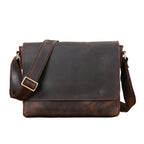 A dark brown distressed leather messenger bag with a wide adjustable shoulder strap and a gold-tone buckle, displayed on a wooden surface with a neutral gray background.