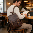 A man sitting at a wooden table in a cozy cafe, reading a book while wearing a dark brown distressed leather backpack with brass hardware.